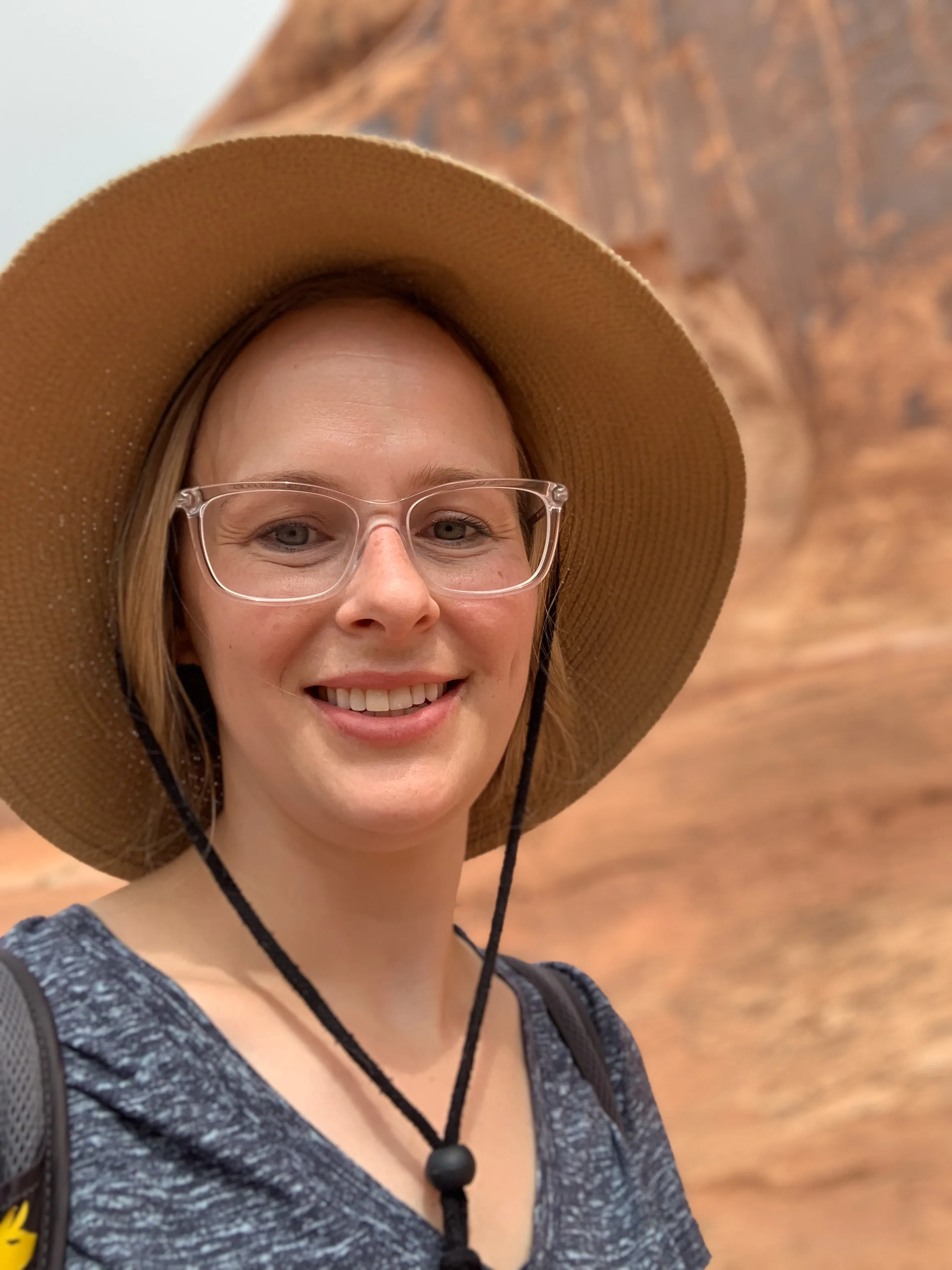 Kerianne Dyer wearing a sun hat and glasses, smiling, in front of a red rock landscape