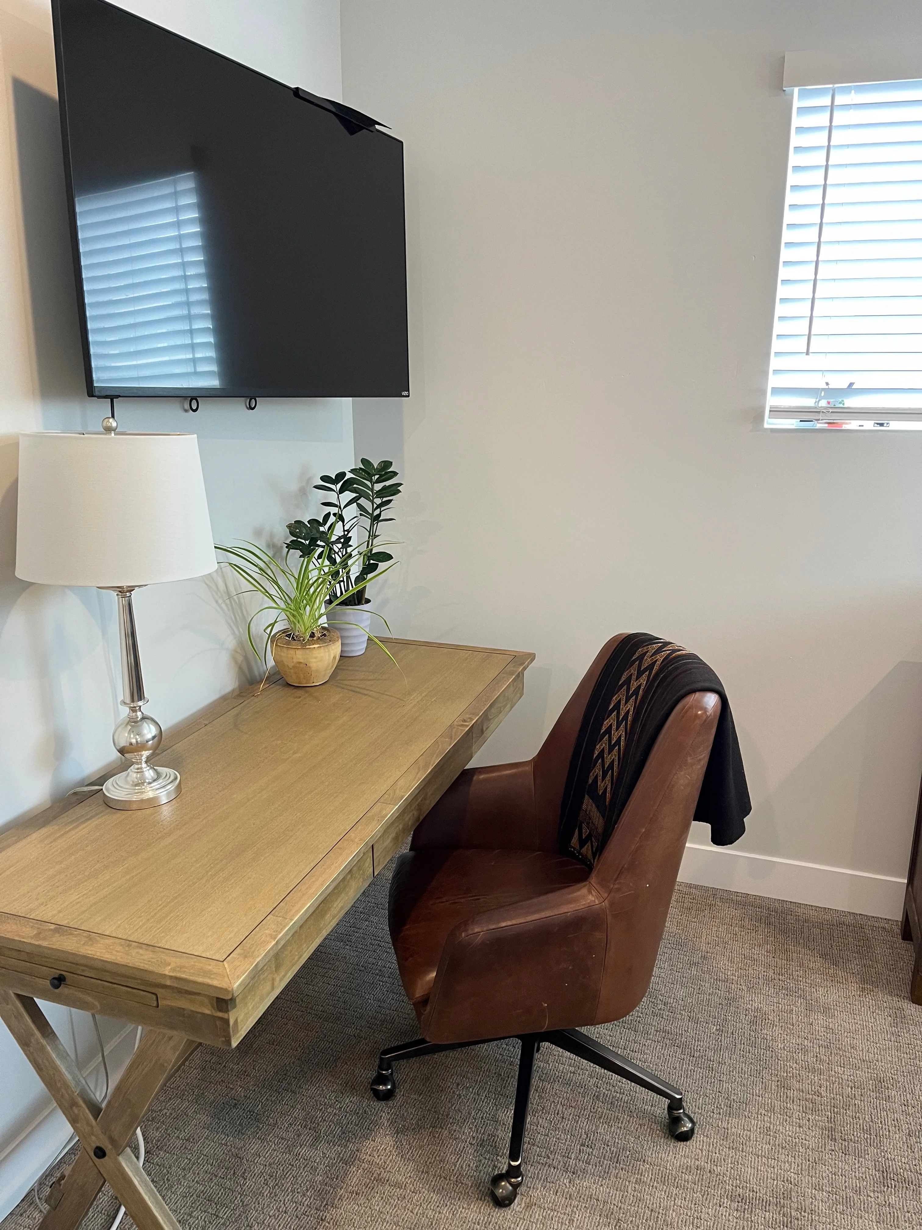 Kerianne's home office: wooden desk, lamp, plant, brown leather chair, mounted screen — where she meets clients from